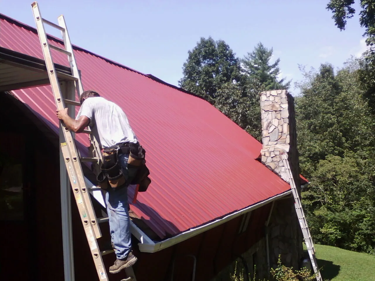 Roofer on a ladder beside a red metal roof and stone chimney — Soffit & Fascia Repair by Roofing Brothers in Sedro Woolley